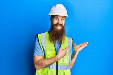 Poster - Redhead man with long beard wearing safety helmet and reflective jacket amazed and smiling to the camera while presenting with hand and pointing with finger.