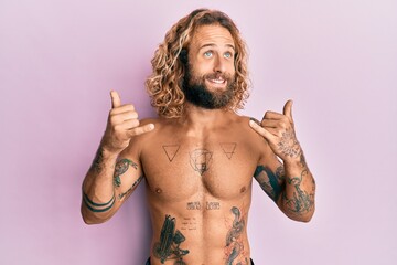 Handsome man with beard and long hair standing shirtless showing tattoos doing shaka sign smiling looking to the side and staring away thinking.