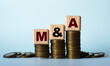 © Zhanna - M and A - acronym on wooden cubes. Which stand on stacks of coins on a light background