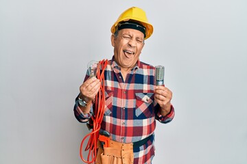 Sticker - Senior hispanic man wearing handyman uniform holding led lightbulb and incandescent bulb sticking tongue out happy with funny expression.