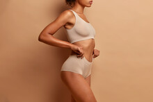 Profile View Of Slim Dark Skinned Woman Dressed In Cropped Top And Panties Demonstrates Her Perfect Body Slender Legs Poses Against Brown Background
