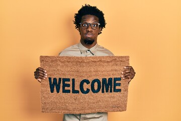 Sticker - Young african american man holding welcome doormat puffing cheeks with funny face. mouth inflated with air, catching air.