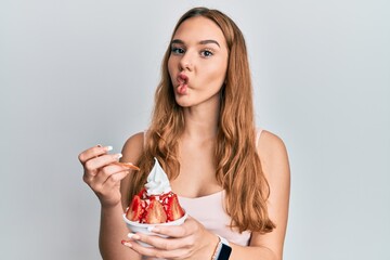 Poster - Young blonde woman eating strawberry ice cream making fish face with mouth and squinting eyes, crazy and comical.