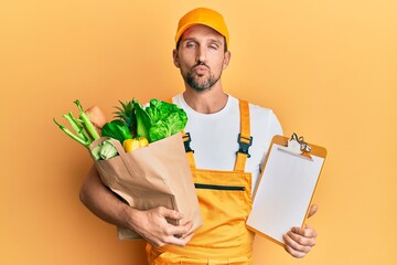 Sticker - Young handsome man wearing courier uniform with groceries and clipboard looking at the camera blowing a kiss being lovely and sexy. love expression.