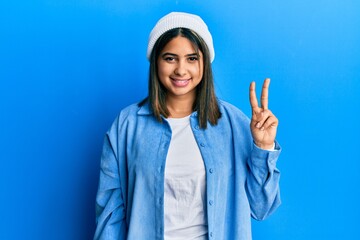 Poster - Young latin woman wearing cute wool cap smiling looking to the camera showing fingers doing victory sign. number two.