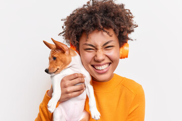 Wall Mural - People animals friendship concept. Overjoyed curly haired woman smiles tenderly holds small pedigree puppy closely to face listens music via wireless headphones isolated over white background