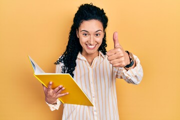 Young hispanic woman with curly hair holding book approving doing positive gesture with hand, thumbs up smiling and happy for success. winner gesture.