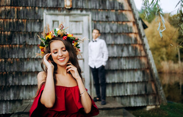 Beautiful girl in a burgundy dress standing near a wooden house. Cute guy on the background