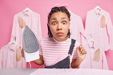 Greatly surprised ethnic woman with braids stares shocked at camera busy ironing at home holds electric irond dressed in casual clothes does daily domestic chores stands against ironed shirts