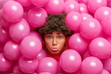 Wall Mural - Displeased curly haired young woman sticks out head through inflated balloons looks sadly at camera expresses negative emotions being in bad mood. Party decorations concept. Spoiled holiday.