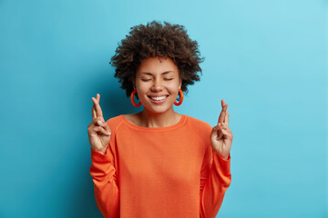 Wall Mural - Lovely cheerful young African American woman with toothy perfect smile keeps fingers crossed believes in good luck wears orange jumper isolated over blue backgound. Superstitious female makes wish