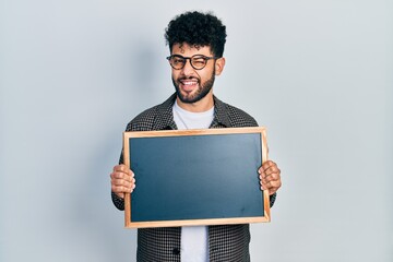 Sticker - Young arab man with beard holding blackboard winking looking at the camera with sexy expression, cheerful and happy face.