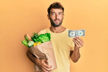 Poster - Handsome man with beard holding groceries and 1 american dollar banknote relaxed with serious expression on face. simple and natural looking at the camera.