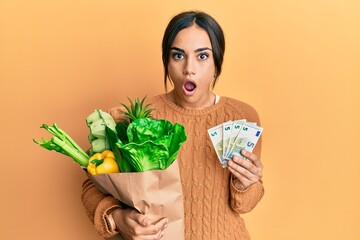 Wall Mural - Young brunette woman holding groceries bag and 5 euros banknotes afraid and shocked with surprise and amazed expression, fear and excited face.