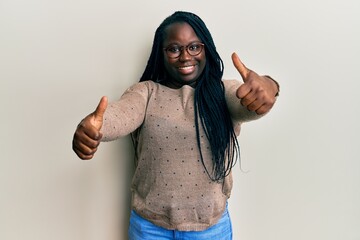 Wall Mural - Young black woman with braids wearing casual clothes and glasses approving doing positive gesture with hand, thumbs up smiling and happy for success. winner gesture.