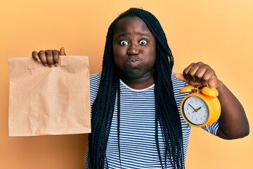 Poster - Young black woman with braids holding take away food and alarm clock puffing cheeks with funny face. mouth inflated with air, catching air.