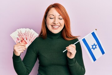 Poster - Beautiful redhead woman holding 20 shekels banknotes and israel flag smiling and laughing hard out loud because funny crazy joke.