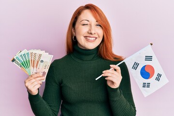 Sticker - Beautiful redhead woman holding south korean won banknotes and flag winking looking at the camera with sexy expression, cheerful and happy face.