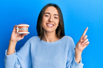 Sticker - Young hispanic girl holding orthodontic smiling happy pointing with hand and finger to the side