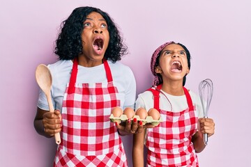 Canvas Print - Beautiful african american mother and daughter cooking cake using baker whisk angry and mad screaming frustrated and furious, shouting with anger looking up.