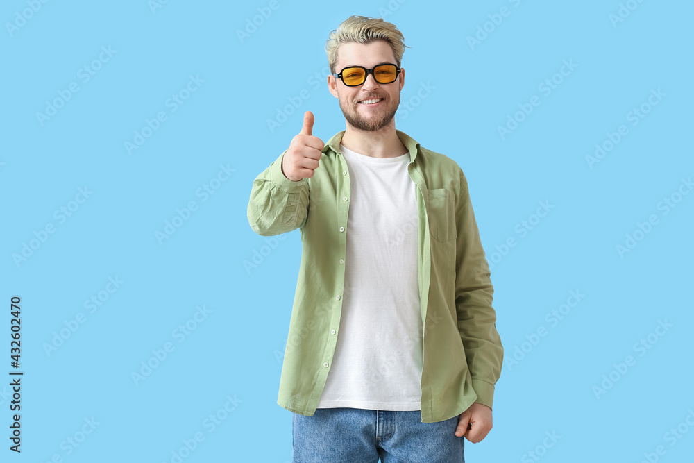 Young man with stylish sunglasses showing thumb-up on color background