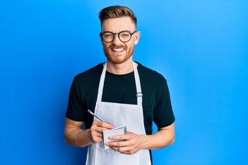 Young redhead man wearing waiter apron taking order smiling and laughing hard out loud because funny crazy joke.