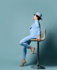 Wall Mural - Full-growth portrait of short haired brunette businesswoman in blue official suit, hat and high-heeled shoes sitting on bar stool over blue background. Stylish female wear and fashion concept