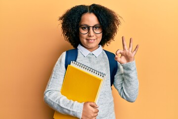 Sticker - Young little girl with afro hair wearing school bag and holding books doing ok sign with fingers, smiling friendly gesturing excellent symbol