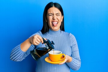 Poster - Beautiful brunette woman holding french coffee maker pouring coffee on cup sticking tongue out happy with funny expression.