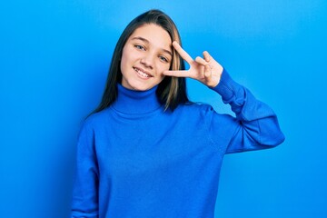 Poster - Young brunette girl wearing turtleneck sweater doing peace symbol with fingers over face, smiling cheerful showing victory