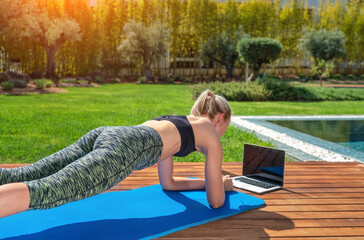 Woman is practicing fitness gymnastics, pilates, on outdoor near house and pool, online lessons with a trainer. With a background to garden.