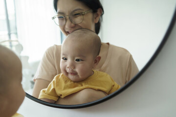 Wall Mural - Cheerful smiling Asian mother and little newborn baby son having fun playing and looking at their faces in mirror at home.