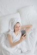 © MyJuly - attractive european woman in a white bed with a towel on her hair chatting on her phone or laptop, relaxing on vacation or traveling, happy white woman in a hotel room chatting with friends online or