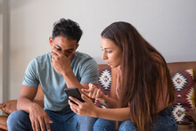 Infidelity. Jealous Girlfriend Showing His Cheating Boyfriend His Phone Demanding Explanation Sitting On Sofa Indoor At Home. Wife Caught Her Husband While Cheating With Mobile Phone - Divorce  People