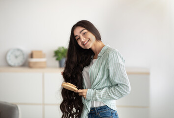 Portrait of lovely Indian lady brushing her wavy long hair, using wooden brush. Domestic spa salon, wellness and beauty