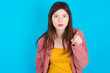 Shocked young beautiful Caucasian woman wearing pink jacket over blue wall points front with index finger at camera and. Surprise and advertisement concept.