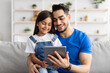 © Prostock-studio - Smiling dad and daughter sitting on couch, using digital tablet