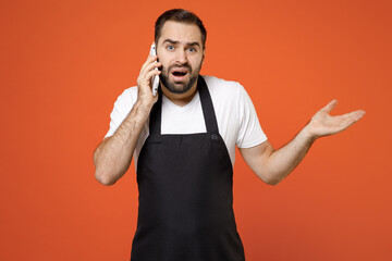 Wall Mural - Young indignant confused man barista bartender barman employee in black apron white t-shirt work in coffee shop talk by mobile phone spread hands isolated on orange background. Small business startup.