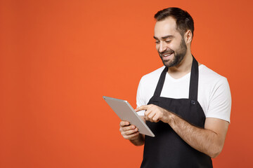 Young smiling fun man 20s barista bartender barman employee in black apron white t-shirt work in coffee shop using tablet pc computer browsing internet isolated on orange background. business startup.