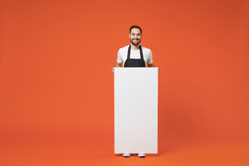 Wall Mural - Full length young man barista bartender barman employee in apron white tshirt work in coffee shop blank sign board promotional content place text isolated on orange background Small business startup