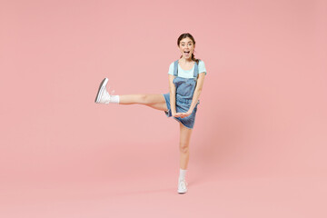 Wall Mural - Full length young smiling joyful happy fun caucasian woman 20s wearing trendy denim clothes blue t-shirt stand with raised up leg isolated on pastel pink background studio portrait. LIfestyle concept.