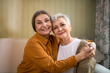 Family, age and mature people concept. Portrait of two joyful elderly sisters with wrinkles and gray hair sitting indoors on comfortable couch, embracing, happy to see each other after lock down