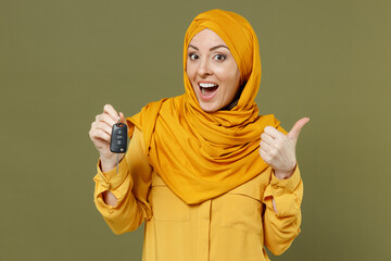 Wall Mural - Smiling fun young arabian asian muslim woman 20s in abaya hijab yellow clothes holding car key fob keyless system show thumb up isolated on olive green background. People uae islam religious concept