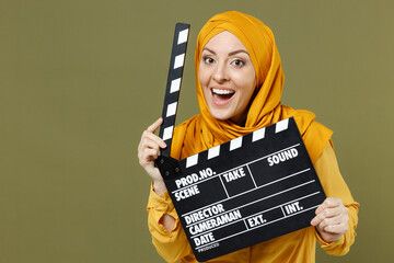 Smiling fun happy young arabian asian muslim woman in abaya hijab yellow clothes holding classic black film making clapperboard isolated on olive green background. People uae islam religious concept