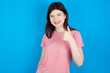 young beautiful Caucasian woman wearing pink T-shirt over blue wall doing happy thumbs up gesture with hand. Approving expression looking at the camera showing success.