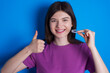 © Roquillo - young beautiful Caucasian woman wearing purple T-shirt over blue wall holding an invisible braces aligner and rising thumb up, recommending this new treatment. Dental healthcare concept.