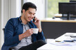 © Bangkok Click Studio - A good looking young businessman sitting and holding a cup of coffee during a morning coffee break while talking on a mobile phone happily in the modern office.