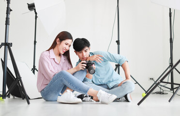 Portrait of two handsome man and beautiful woman wearing casual shirts, jeans, checking photos in camera while sitting on floor in photo studio with white background cutout and lighting equipment.