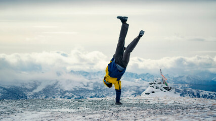 A guy stands on one arm in the snowy mountains in winter. Bright jacket. above the clouds