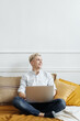 © diignat - Young attractive smiling blonde guy is browsing at his laptop, sitting at home on the cozy sofa at home, wearing casual outfit